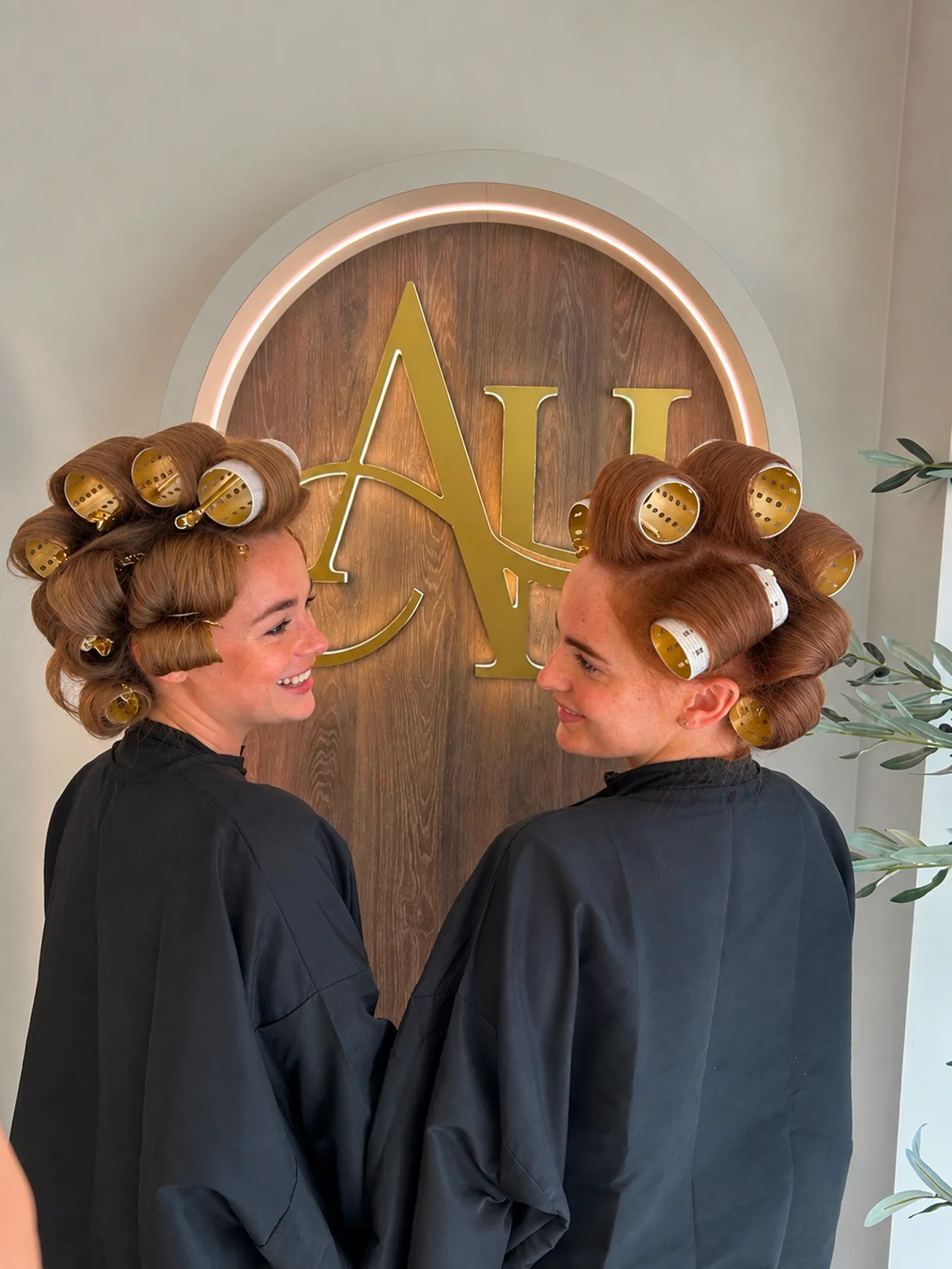 Two women wearing hair rollers smile brightly, showcasing a joyful moment together